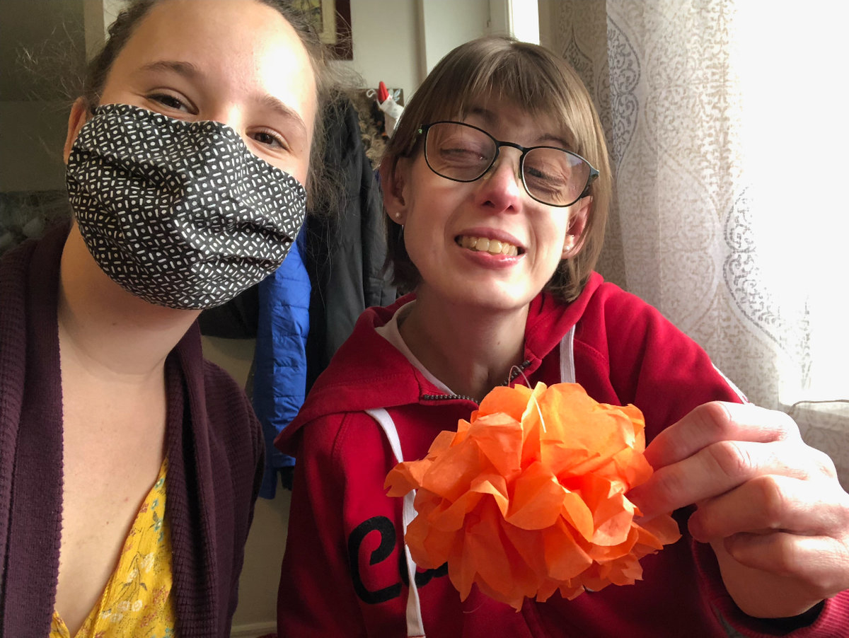 Stephanie and with a Personal Support Worker smiling and holding up an orange tissue paper flower that she made.