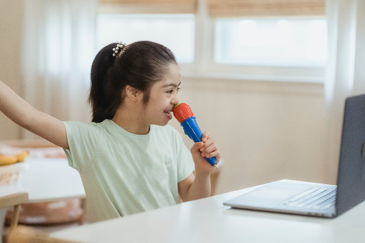 A young girl with a disability sits at a desk with a laptop computer on it. The desk is in a bedroom with a bed and open window behind her. She holds one arm up, and in her other hand she holds a toy microphone to her mouth. It looks like she is singing along with or singing to someone on the computer. 